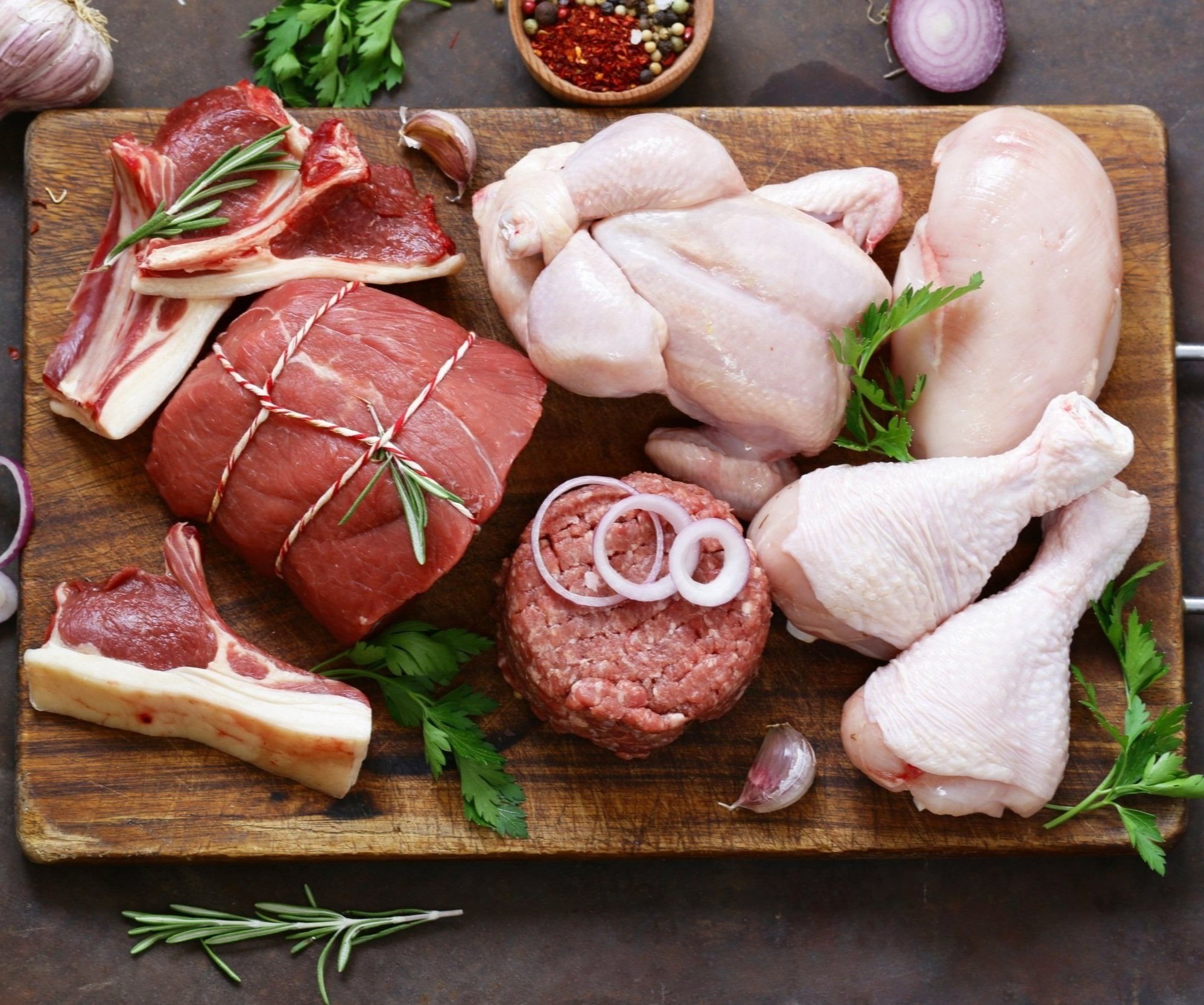 Assorted raw meats on a wooden cutting board, including beef cuts, chicken breasts, drumsticks, and ground beef, garnished with fresh herbs like rosemary and parsley, with garlic and red onion nearby.