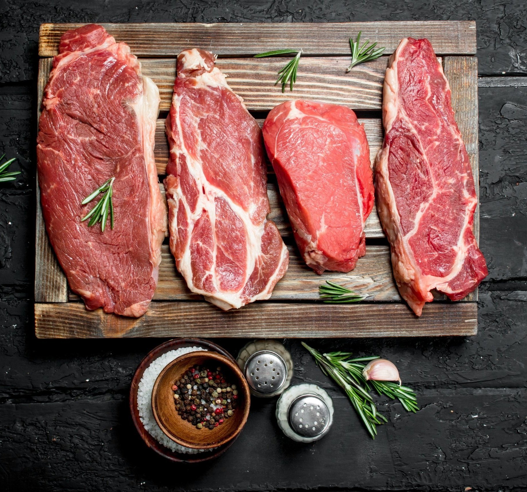Assorted cuts of raw beef steaks displayed on a wooden cutting board, garnished with fresh rosemary, accompanied by a bowl of mixed peppercorns, salt shakers, and a garlic clove, set against a dark textured background.