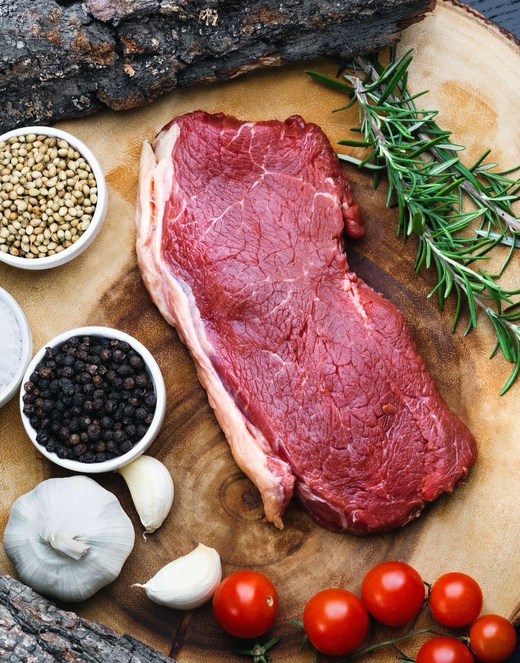 Fresh raw steak surrounded by garlic, cherry tomatoes, black pepper, coriander seeds, sea salt, and sprigs of rosemary on a wooden cutting board. Perfect for grilling or roasting.